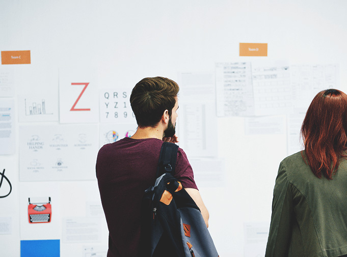 Two people viewing design research papers and sketches pinned to a white studio wall