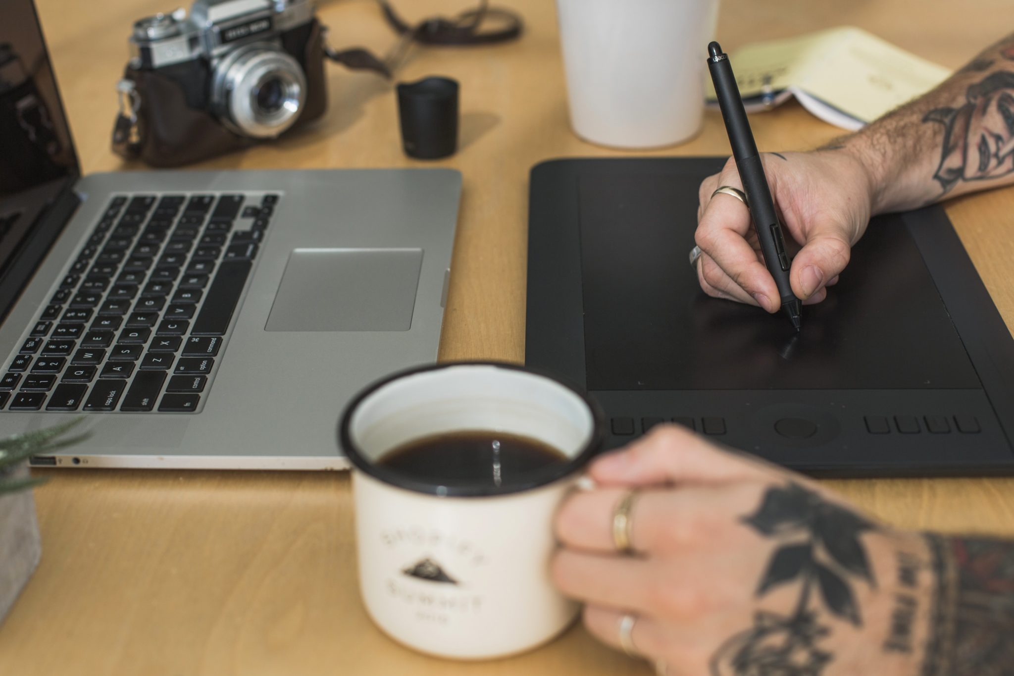 Designer using a stylus on a graphics tablet beside a laptop, camera, and coffee mug