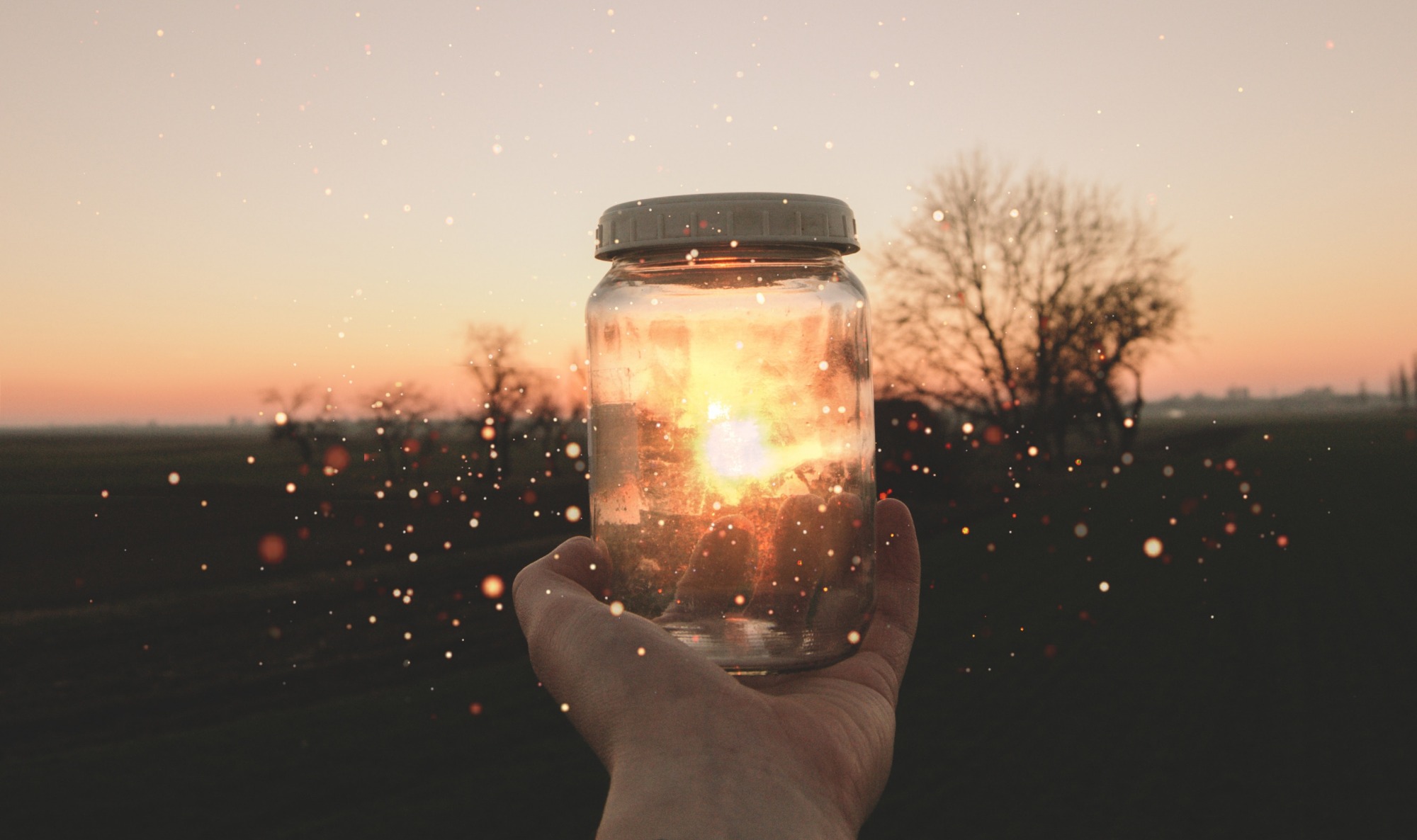 Hand holding a glass jar with glowing sunset light inside at dusk in a field